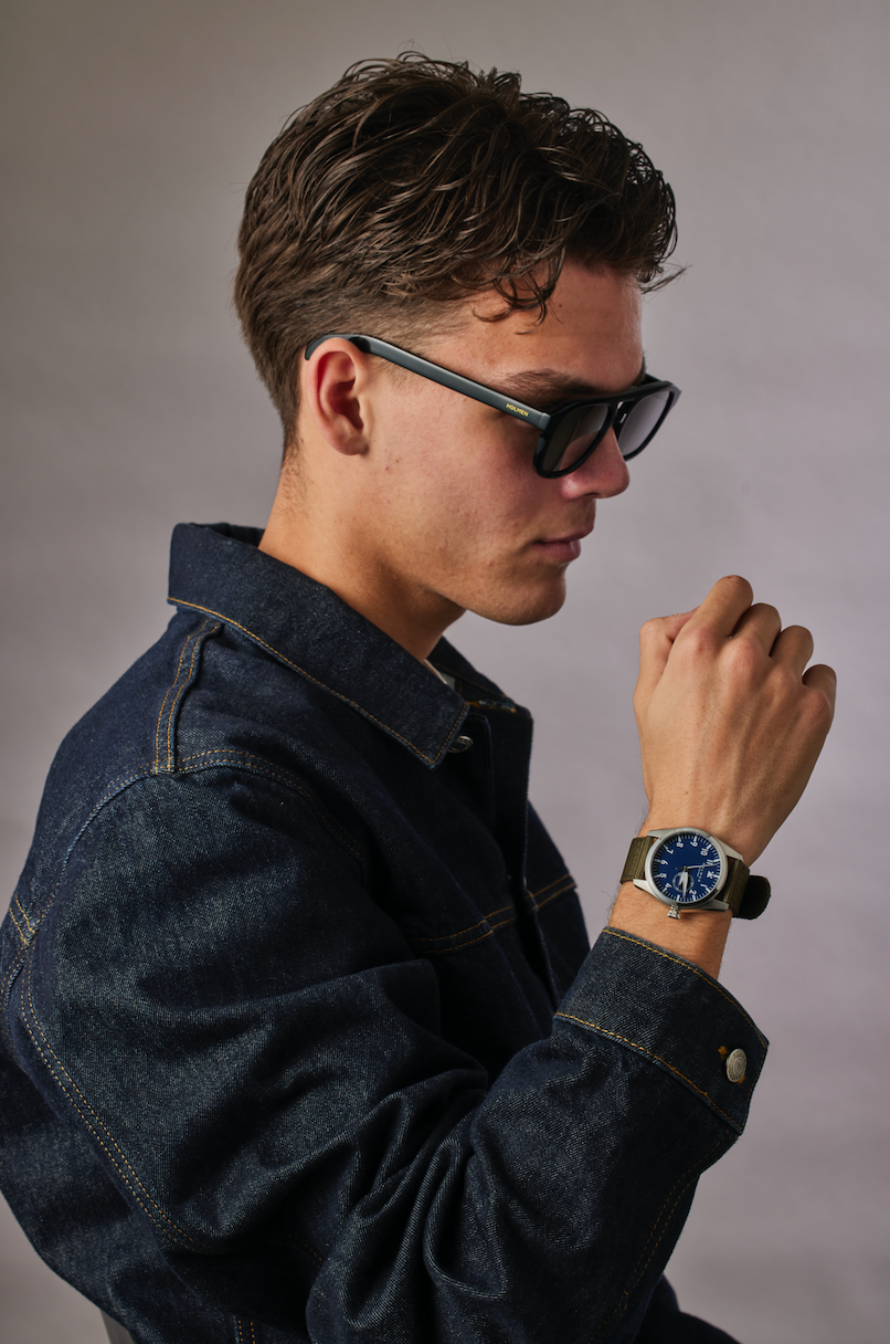 Man wearing a denim jacket and sunglasses, looking at his watch against a gray background