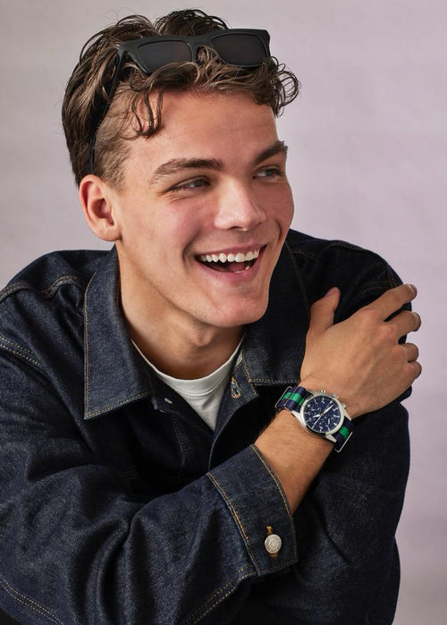 Man wearing a denim jacket and a watch, with sunglasses on his head, against a plain background