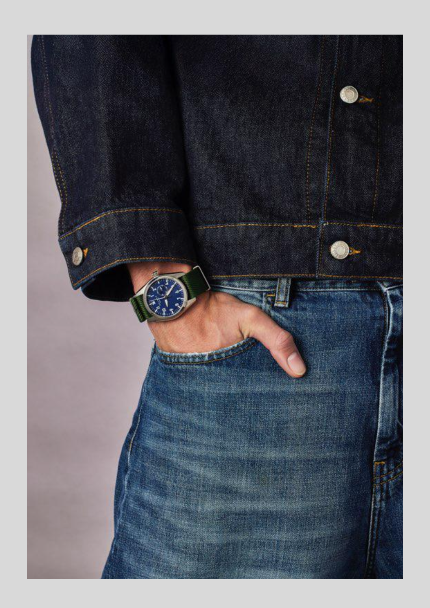 Person wearing a blue denim jacket and jeans with a watch on a neutral background