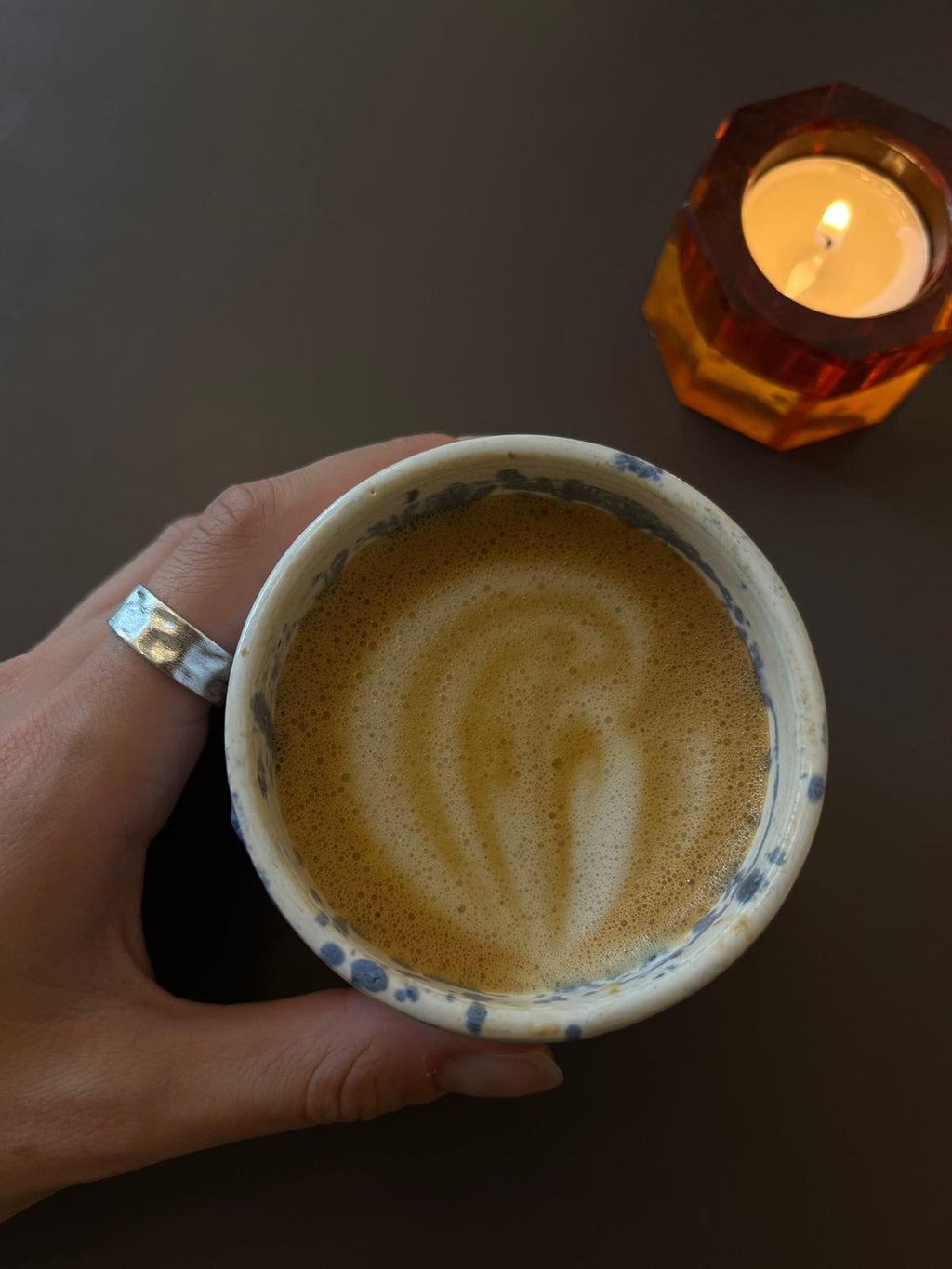 Hand holding a cup of coffee with latte art next to a lit candle on a dark surface